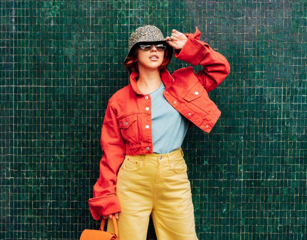 Person standing against a green tiled wall, wearing a red jacket, yellow pants, grey shirt, animal print bucket hat, sunglasses, and holding an orange bag.