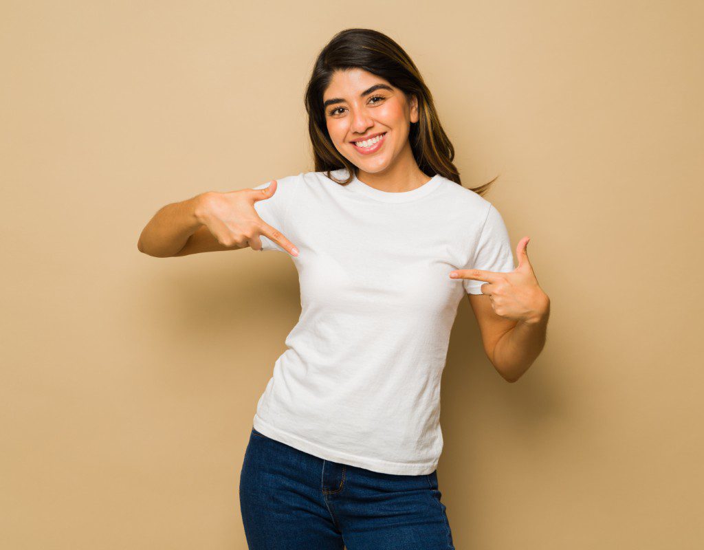 A young woman wearing a plain white t-shirt and jeans smiles and points at her shirt while standing in front of a beige background.