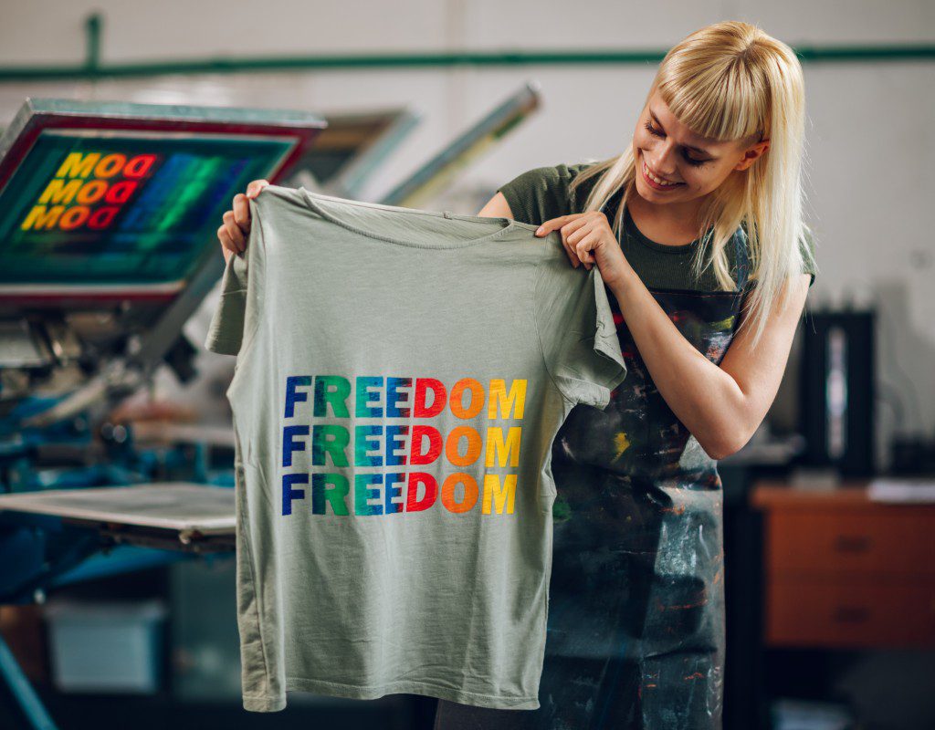 A woman holds up a T-shirt printed with the word "FREEDOM" in blue, green, red, and yellow letters in a screen-printing workshop.