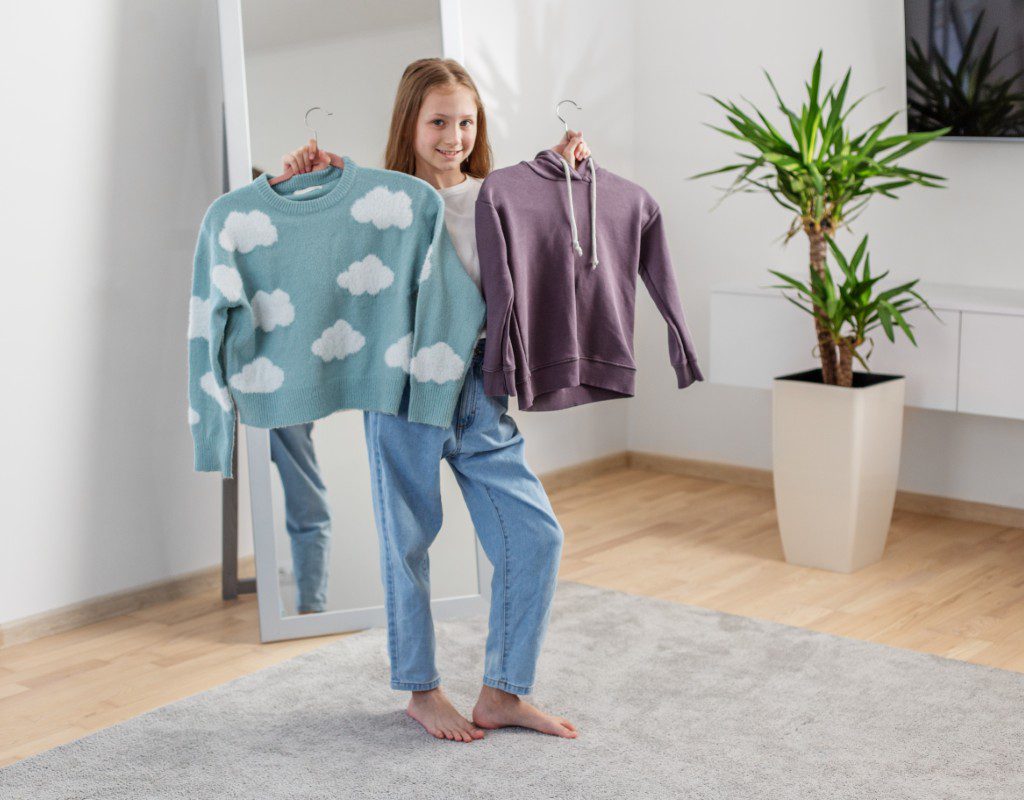 A girl standing barefoot in a room holds up a blue sweater with cloud patterns in one hand and a purple hoodie in the other. A large plant and a mirror are in the background.