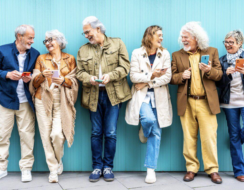 Six older adults stand in a row against a blue wall, smiling and chatting while holding smartphones.